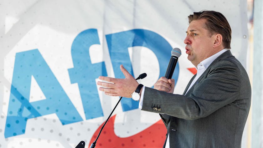 Maximilian Krah auf einer Bühne der Kundgebung in Dresden. Er hält ein Mikrofon an seinen Mund und gestikuliert mit der linken Hand. Im Hintergrund eine Flagge mit AFD-Logo.&nbsp;