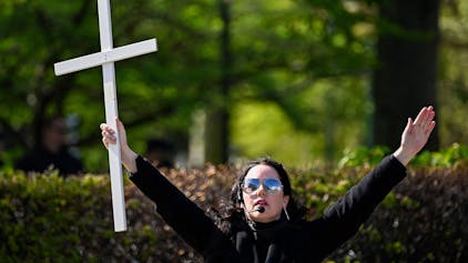 Eine Frau mit Sonnenbrille und Kopfmikrofon breitet die Arme weit aus. In ihrer rechten Hand hält sie ein Kreuz.&nbsp;