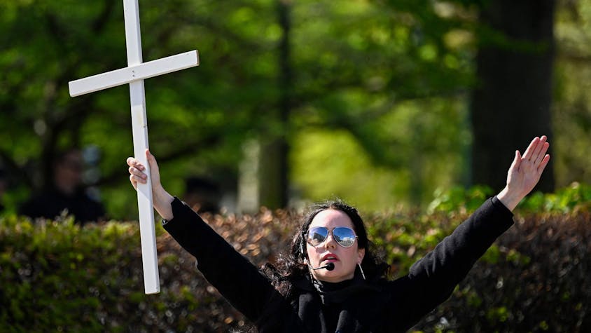 Eine Frau mit Sonnenbrille und Kopfmikrofon breitet die Arme weit aus. In ihrer rechten Hand hält sie ein Kreuz.&nbsp;