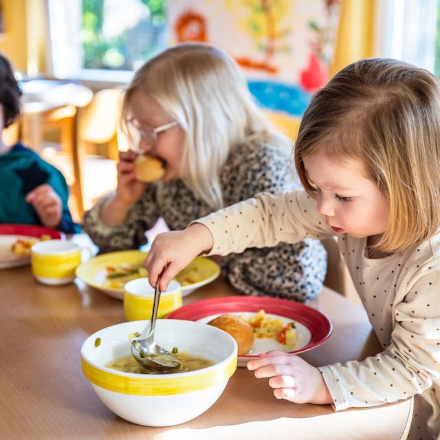 Ein Mädchen in einer Kita füllt mit einer Kelle Gemüsesuppe auf ihren Teller. Zwei weitere Kinder sitzen im Hintergrund am Tisch.&nbsp;