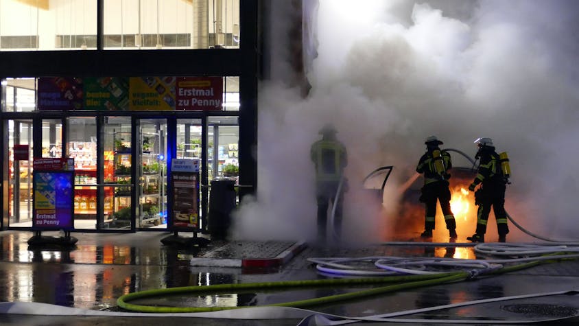Feuerwehrleute löschen ein brennendes Auto nebem dem Eingang eines Supermarkts.