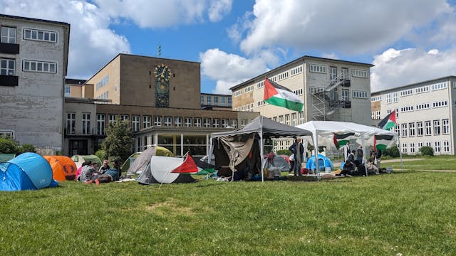 Zelte und ein Pavillon mit Palästina-Flaggen stehen auf der Uniwiese, im Hintergrund ist das Hauptgebäude der Uni Köln zu sehen.