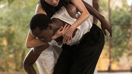 Auf dem Bild sind ein Tänzer und eine T#nzerin zu sehen, in schwarz-weißer Kleidung. Die Tänzerin umklammert den Tänzer mit ihren Händen, er ist in geduckter Haltung.&nbsp;