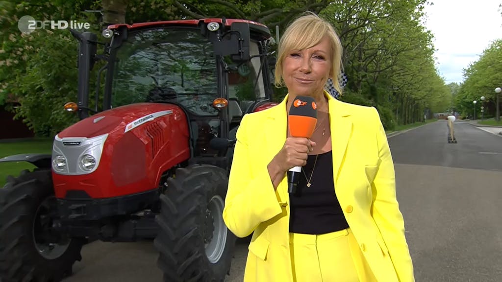 Hier zu sehen: Moderatorin Andrea Kiewel in knallgelbem Anzug, vor einem Traktor bei einer Challenge des ZDF-„Fernsehgarten“ stehend.