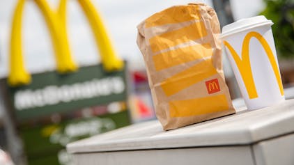 Eine McDonald's To-Go-Tüte und ein To-Go-Becher stehen vor einer McDonald's-Filiale.