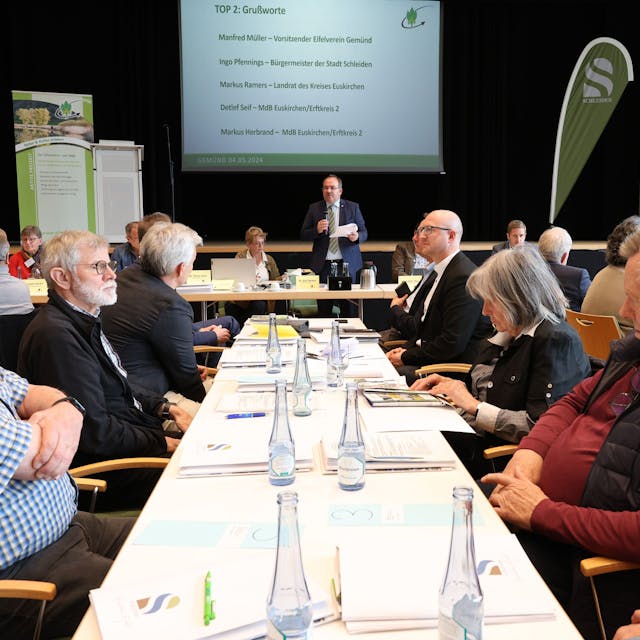 Zahlreiche Personen sitzen bei der Tagung des Eifelvereins im Kursaal Gemünd an Tischen. In der Mitte steht Hauptvorsitzender Dr. Ralf Nolten.