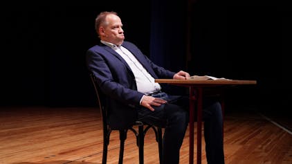 Kabarettist Thomas Schreckenberger sitzt im Stadttheater in Euskirchen an einem Tisch. Er gibt eine gelungene Version von Klaus Kinski zum besten.