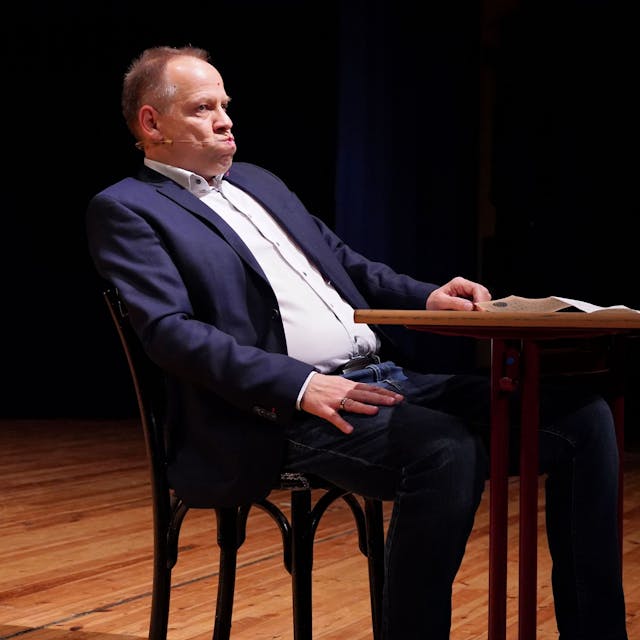 Kabarettist Thomas Schreckenberger sitzt im Stadttheater in Euskirchen an einem Tisch. Er gibt eine gelungene Version von Klaus Kinski zum besten.