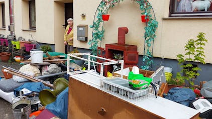 Eine Frau steht an ihrer Haustür. Vor dem Haus stapeln sich Möbel und weitere Gegenstände, die beim Hochwasser in Dahlem zerstört wurden.