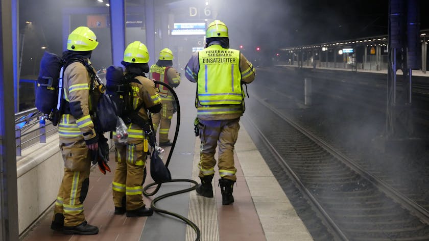 Feuerwehrleute stehen am Bahnsteig, unter dem Rauch hervordringt.