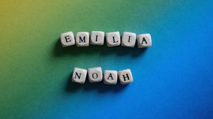 Holzwürfel mit Buchstaben, die die Namen Emilia und Noah ergeben, liegen auf einer bunten Pappe auf einem Tisch.&nbsp;