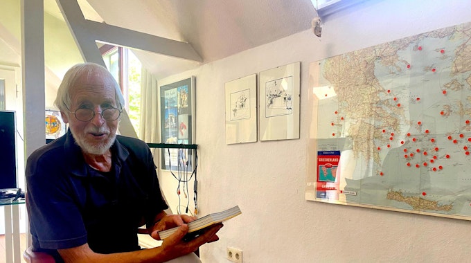 Kurt Schreiner vor einer Weltkarte mit vielen Pins an den bereisten Orten. In der Hand hält er eines von seinen selbstgeschriebenen Reisebüchern.