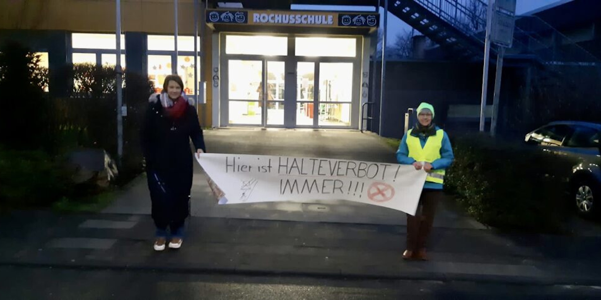Ute Arnold (l.) und Anne Keller mit einem Banner vor der Rochusschule. Die Verkehrssituation dort ist angespannt.