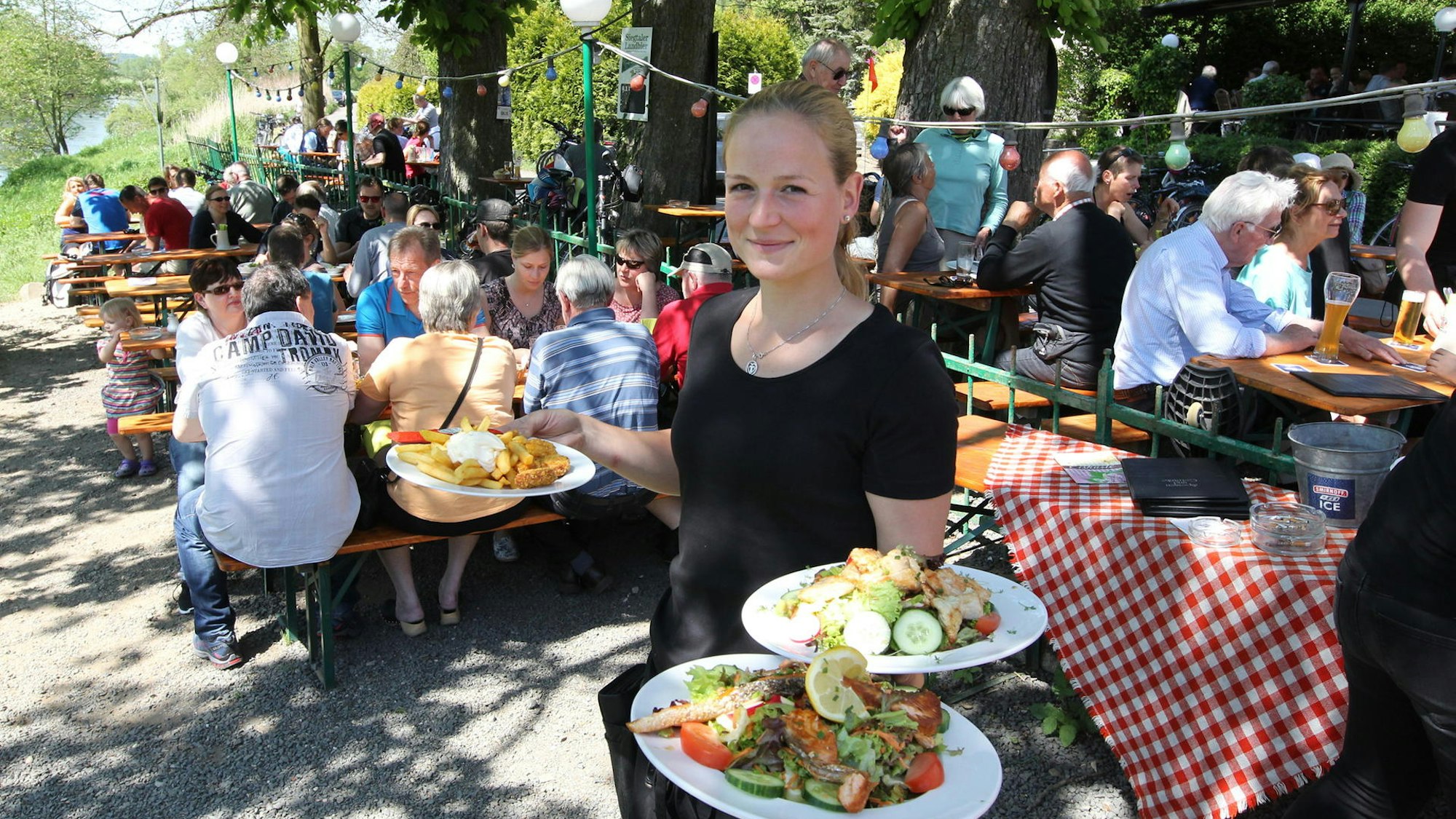 Eine Frau hält drei Teller mit Gerichten in der Hand. Im Hintergrund stehen vollbesetzte Tische unter Bäumen,