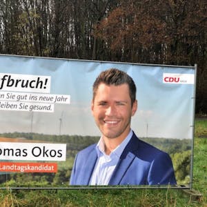 Die CDU hat in Hürth Bauzaunbanner des Landtagskandidaten Thomas Okos aufgestellt, der Neujahrsgrüße überbringt.