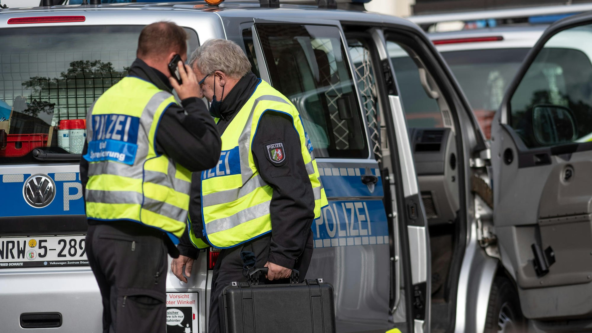 Ein Polizist steht telefonierend an einem Tatort, sein Kollege geht mit Spurensicherungskoffer zum Einsatzwagen.