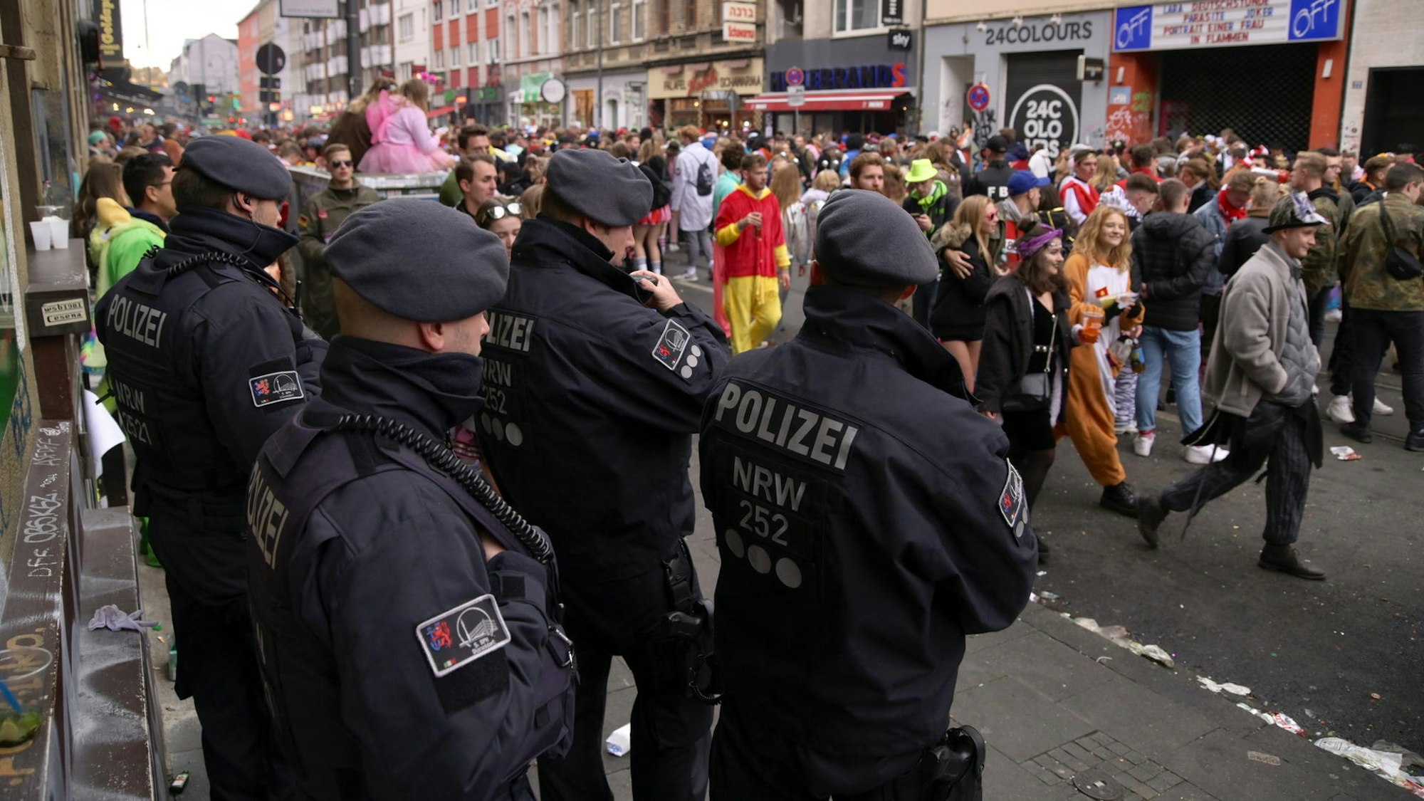 Polizisten einer Hundertschaft stehen am Rand der Zülpicher Straße und beobachten kostümierte Menschen, die vorüberziehen.