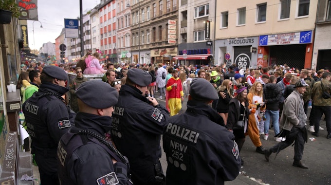 Polizisten einer Hundertschaft stehen am Rand der Zülpicher Straße und beobachten kostümierte Menschen, die vorüberziehen.