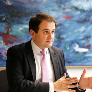 NRW-Europaminister Nathanael Liminski (CDU) in einer Gesprächssituation, den Gesprächspartner sieht man nicht. Er sitzt vor einem großen abstrakten Gemälde mit viel Blau, trägt einen dunklen Anzug, weißes Hemd und rosafarbene Krawatte.