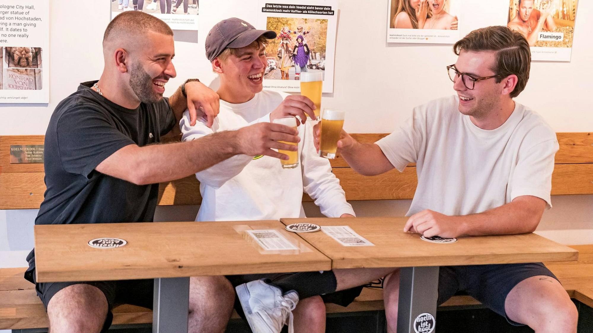 Firat Mercan, Tim Schmitz und Julius Kahleis sitzen an einem Tisch und stoßen lachend mit Bier an.