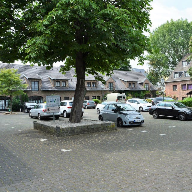 Ein gepflasterter Platz mit einigen eingefassten Bäumen und daneben geparkte Autos.&nbsp;