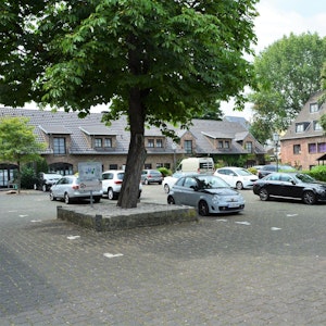 Ein gepflasterter Platz mit einigen eingefassten Bäumen und daneben geparkte Autos.
