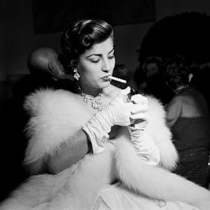 Irene Papas mit Zigarette auf einer Party. Sie trägt Pelz und weiße Handschuhe.