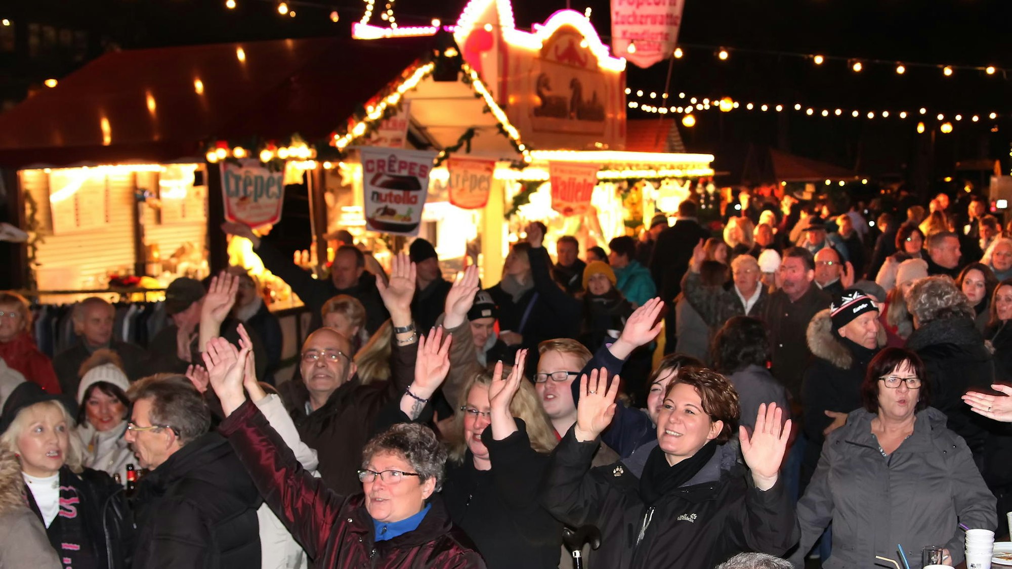 Viele Menschen stehen vor einer Bude auf dem Weihnachtsmarkt und strecken die Hände in die Luft.