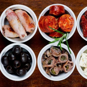 Spanische Tapas, wie hier zum Beispiel Datteln in Speck, Chorizo, getrocknete Tomaten, Oliven, Sardellen und Schafskäse (von oben links nach unten rechts, Symbolbild))