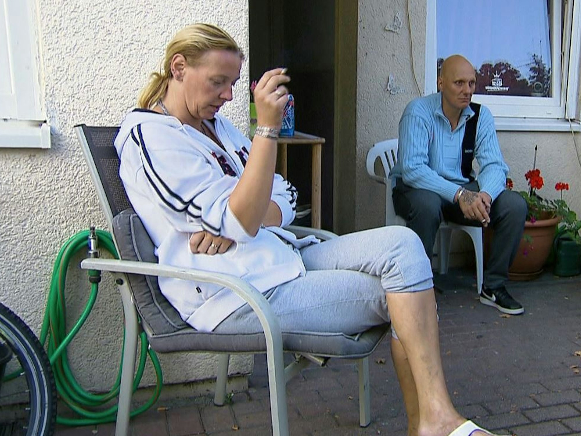 Katrin sitzt auf einem Stuhl auf der Terrasse und raucht eine Zigarette. Im Hintergrund sitzt Ehemann Elvis auf einem Stuhl.