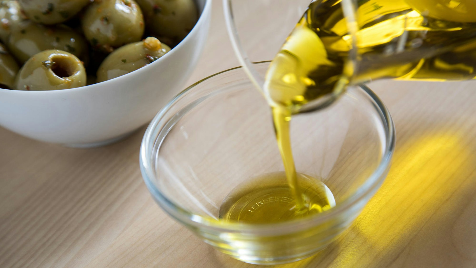 Aus einer Glaskaraffe wird Olivenöl in eine Glasschale gegossen, im Hintergrund eine Porzellanschale mit grünen Oliven.