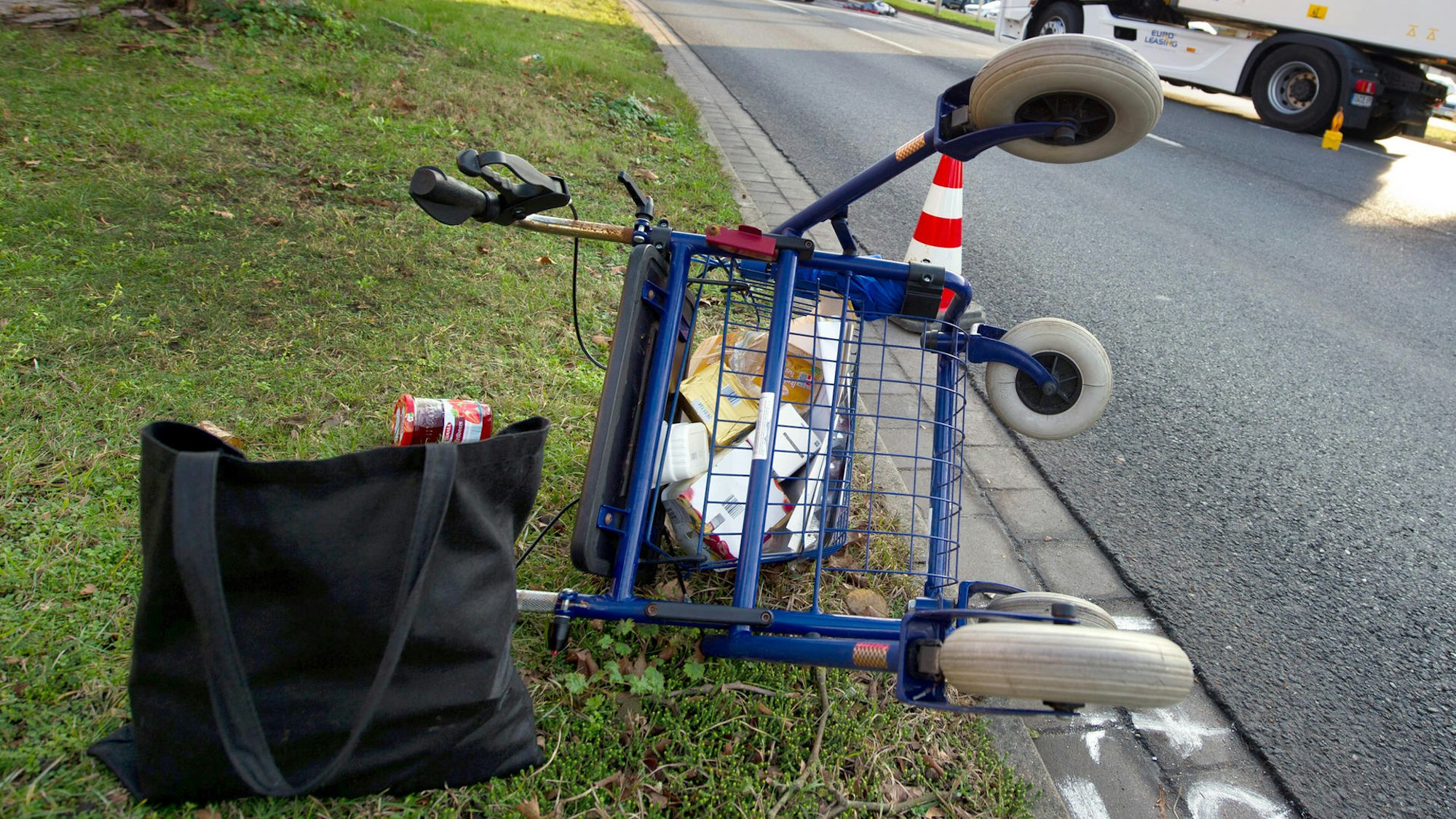 Ein Rollator liegt umgekippt auf einem Grünstreifen neben einer Straße. Daneben steht eine Einkaufstasche.