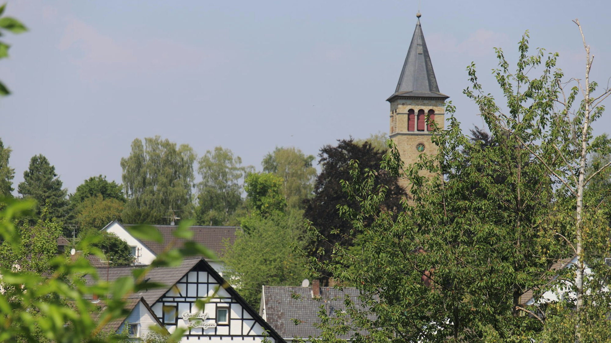 Das Foto zeigt einen Kirchturm und Fachwerkhäuser hinter Baumwipfeln.