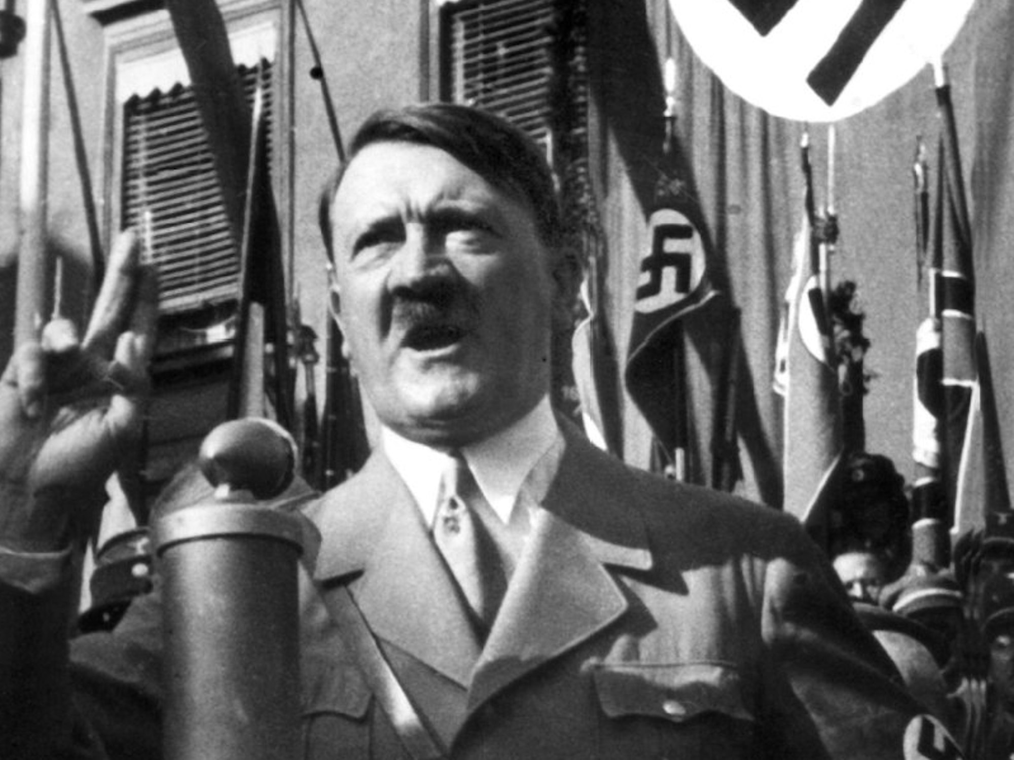 Adolf Hitler hält eine Rede. Im Hintergrund wehen Hakenkreuz-Fahnen.