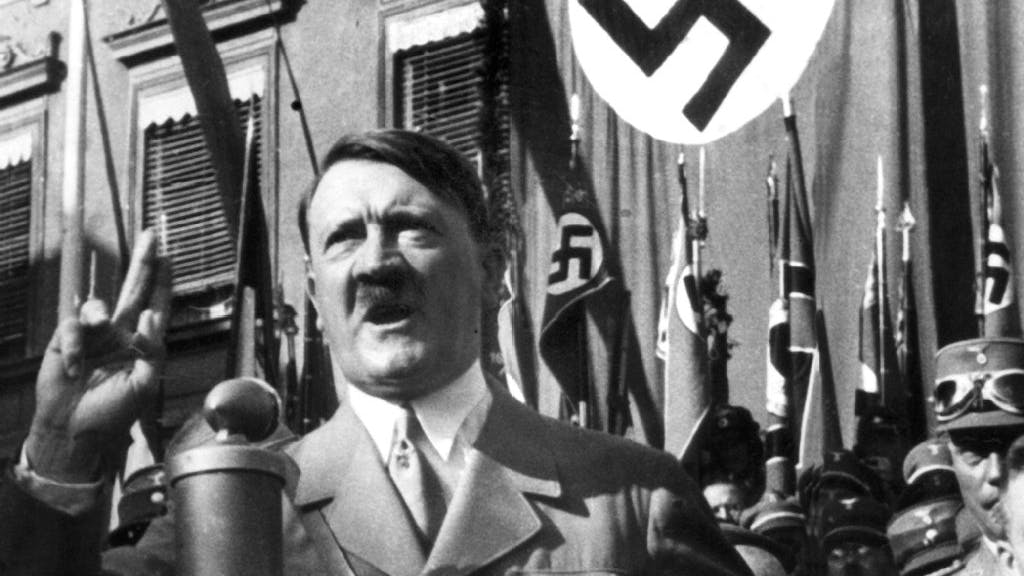 Adolf Hitler hält eine Rede. Im Hintergrund wehen Hakenkreuz-Fahnen.