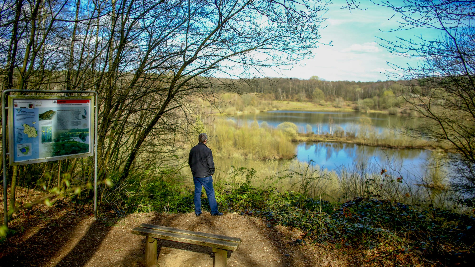 Mann steht an einem Rand eines Gewässers, hinter ihm eine Infotafel und eine Holzbank