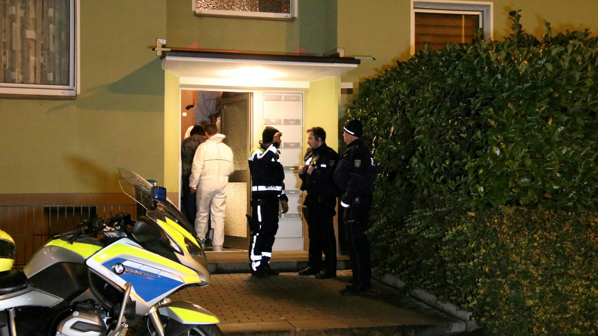 Drei Polizisten stehen vor einem Mehrfamilienhaus, ein Beamter der Spurensicherung in einem Schutzanzug geht in das Haus.