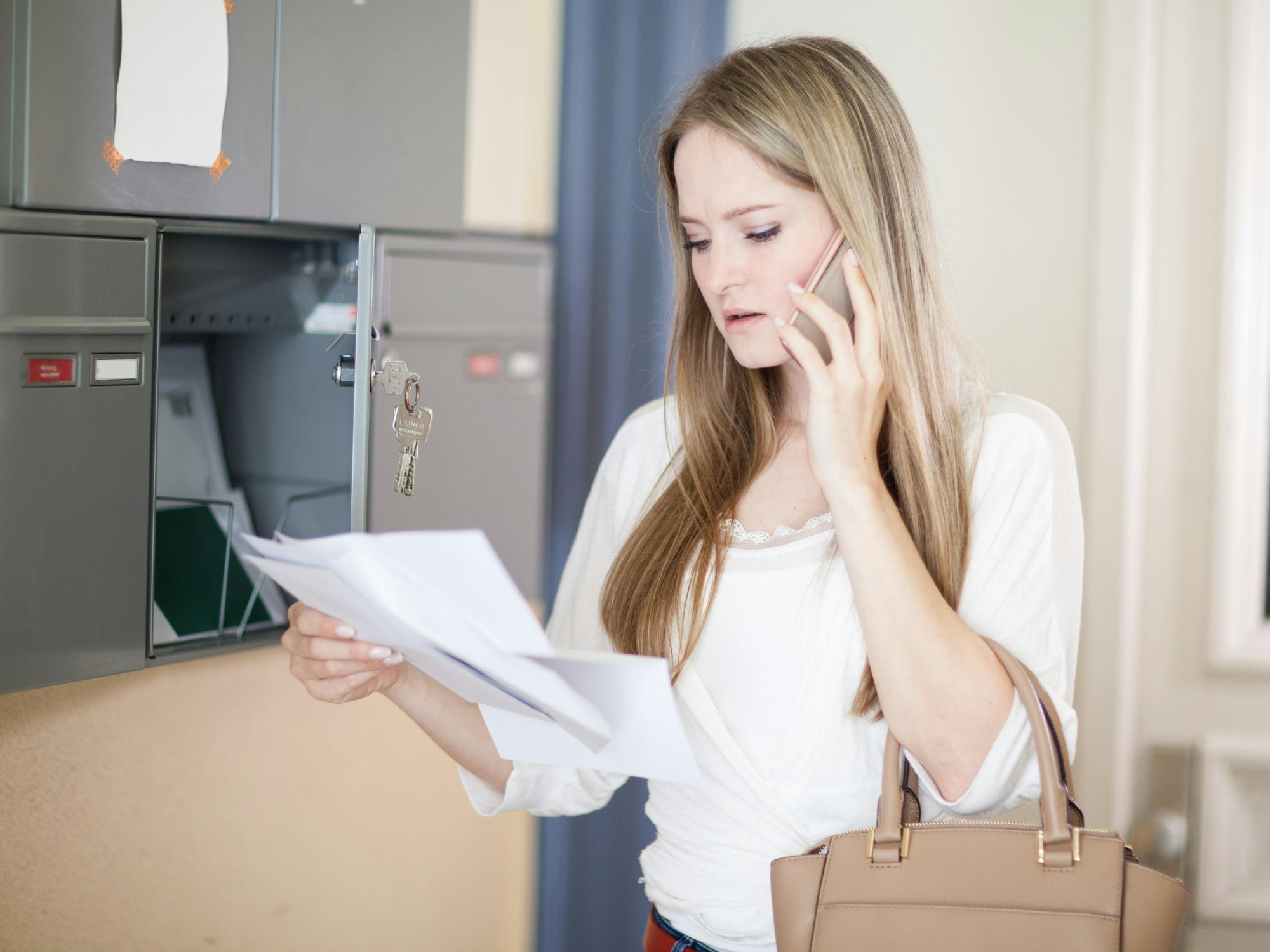 Eine steht mit einem Brief in der Hand vor einem Briefkasten und hält ihr Telefon an ihr Ohr.