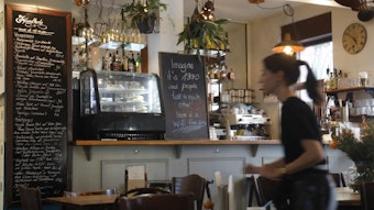Das Café Sehnsucht in Köln von innen, mit zwei Tafeln mit dem aktuellen Angebot und einer geblurrten Thekenkraft