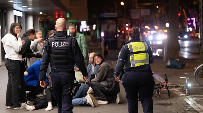 Menschen hocken nachts auf dem Hohenzollernring im Kreis um einen Verletzten, der niedergeschlagen wurde, zwei Polizisten eilen hinzu.