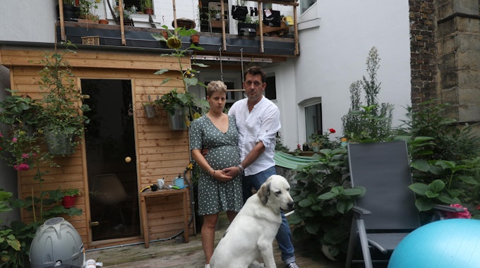Eine schwangere Frau steht mit einem Mann und Hund auf einer Terrasse.