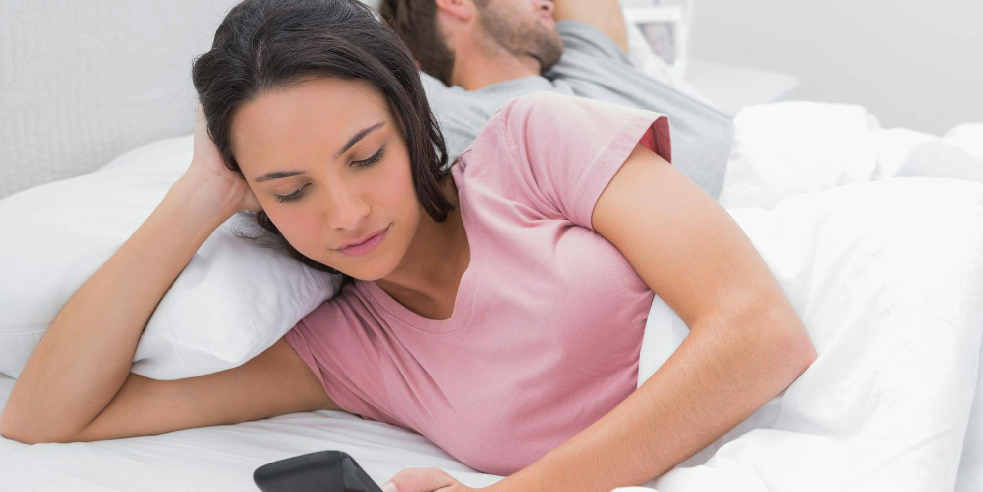 Paar liegt im Bett und Frau schreibt heimlich am Smartphone