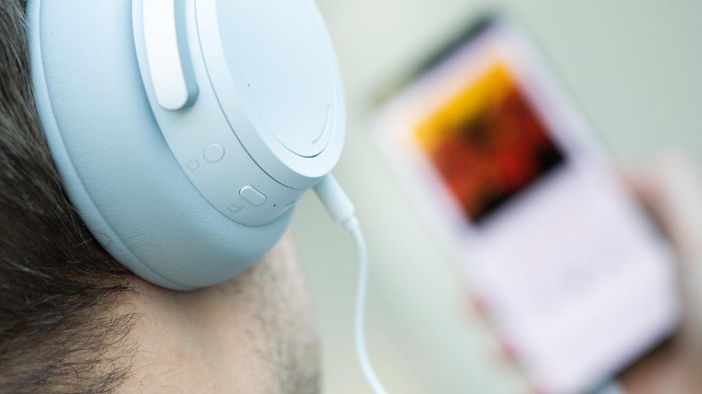 Weißer Over-Ear-Kopfhörer, im Hintergrund unscharf ein Smartphone mit Musikstreamingdienst.
