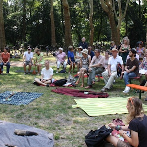 Rund um eine Reihe ausgebreiterte Picknick-Decken sitzen Menschen im Kreis, im Hintergrund eine Baumreihe. Eine Frau redet in ein Mikrofon.