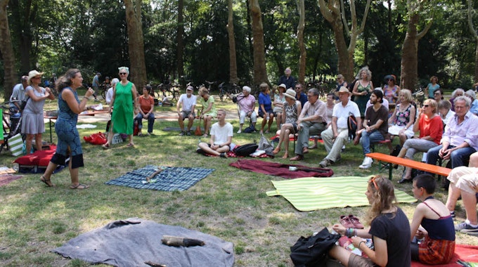 Rund um eine Reihe ausgebreiterte Picknick-Decken sitzen Menschen im Kreis, im Hintergrund eine Baumreihe. Eine Frau redet in ein Mikrofon.