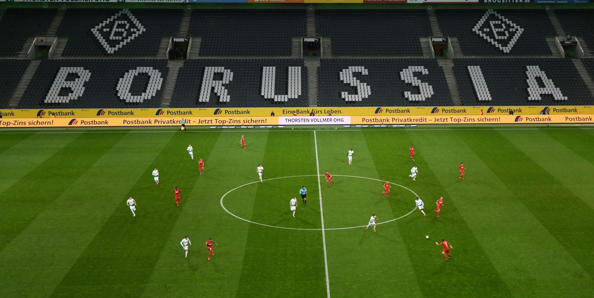 Großes, leeres Stadion, dazu 22 Spieler des 1. FC Köln (in rot) und von Borussia Mönchengladbach, die ein Bundesliga-Spiel austragen.