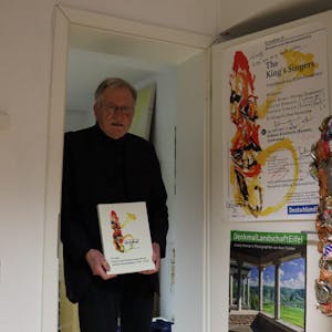 Sein Buch über die spannende Konzertreihe „Grundton D“ hält Wolf Werth in Händen. An der Tür zu seinem Arbeitszimmer hängt noch ein Originalplakat neben einer Figur der Trash People des Künstlers HA Schult.