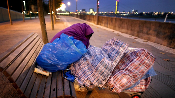 Ein obdachloser Mann sitzt mit einer Decke auf einer Parkbank.