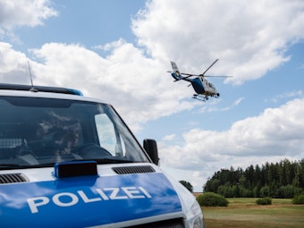 Ein Polizeifahrzeug steht auf einer ländlichen Straße, im Hintergrund fliegt ein Hubschrauber.