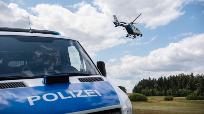 Ein Polizeifahrzeug steht auf einer ländlichen Straße, im Hintergrund fliegt ein Hubschrauber.